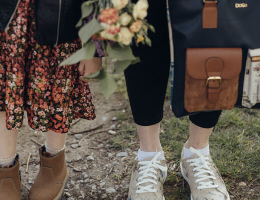 Mutter und Tochter stehen nebeneinander auf einer Wiese. Man sieht nur die Beine. Mutter hält Blumenstrauß und eine Tasche von DOGO in den Händen.