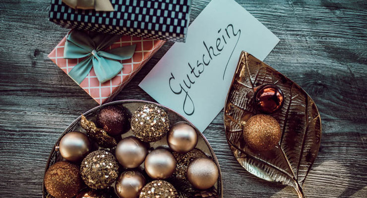 Ein Umschlag mit der Aufschrift "Gutschein" liegt neben Geschenken und Weihnachtskugeln.