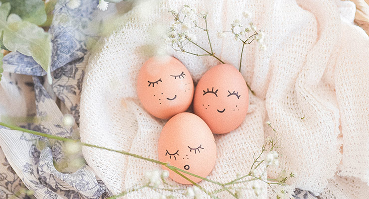 Eier mit aufgemalten Gesichtern auf einem Tuch umgeben von zarten Frühlingsblumen