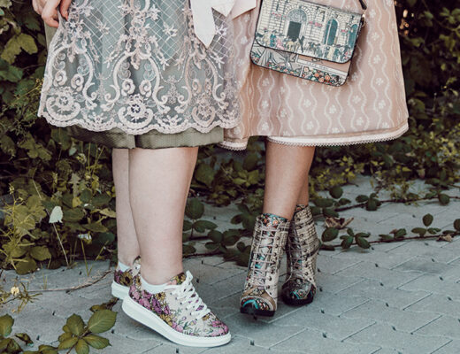 Zwei Frauen im Dirndl stehen vor einem grünen Busch. Man sieht lediglich ihre Rocksäume und ihre Beine. An den Füßen trägt eine von ihnen DOGO Myra Sneaker mit Blumenmotiv und die andere DOGO Chloe mit hohem Absatz und einem Design, dass ein Café in Wien zeigt.