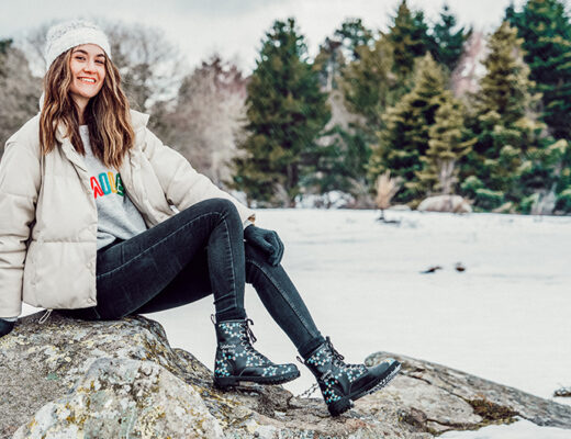 Frau sitzt auf einem Felsen inmitten einer Schneelandschaft. Sie trägt eine helle Jacke, eine dunkle Jeans und DOGO Boots im Design "Celebrate the Day".